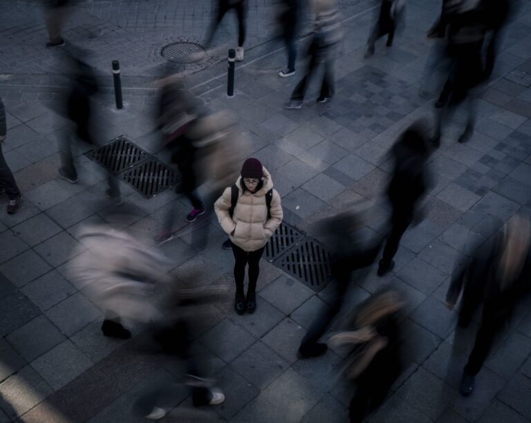 A student stands still in a busy hall. Dozens of people around her are blurred, indicating motion. She appears alone in the crowd.