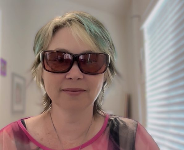 Kim Ashbourne, white, 50ish, with short hair that is a mix of blonde, brown and teal. She wears dark glasses that filter light and sits in her home office.